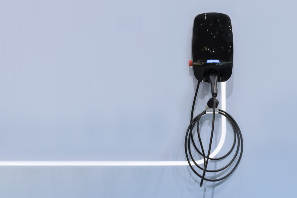 A black wall-mounted electric vehicle charger with a coiled cable against a light gray background.